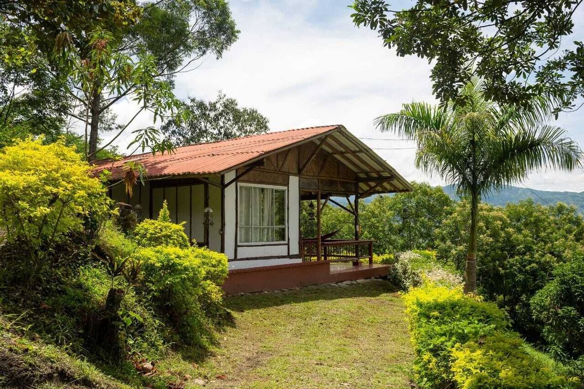 A small, rustic house with a red roof, surrounded by lush greenery and a clear view of distant mountains.