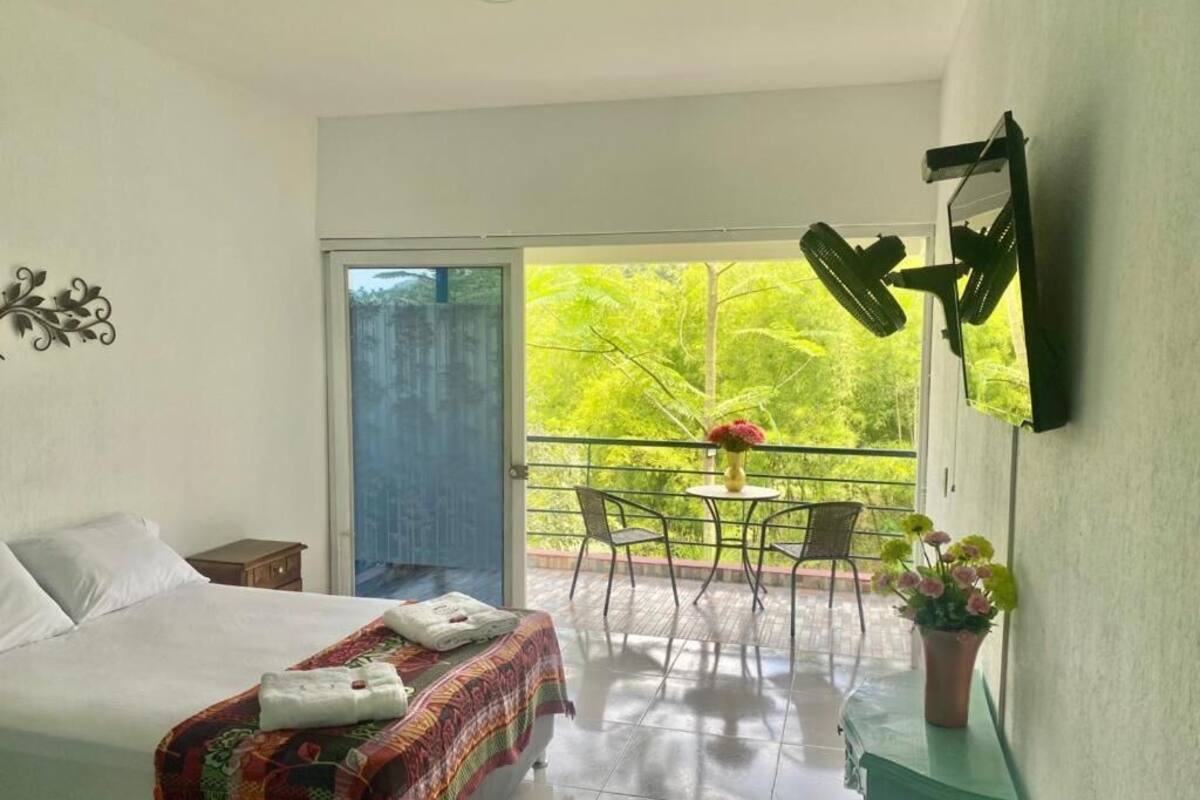 A bedroom with a bed, a bedside table, a balcony with a table and chairs, and a potted plant.