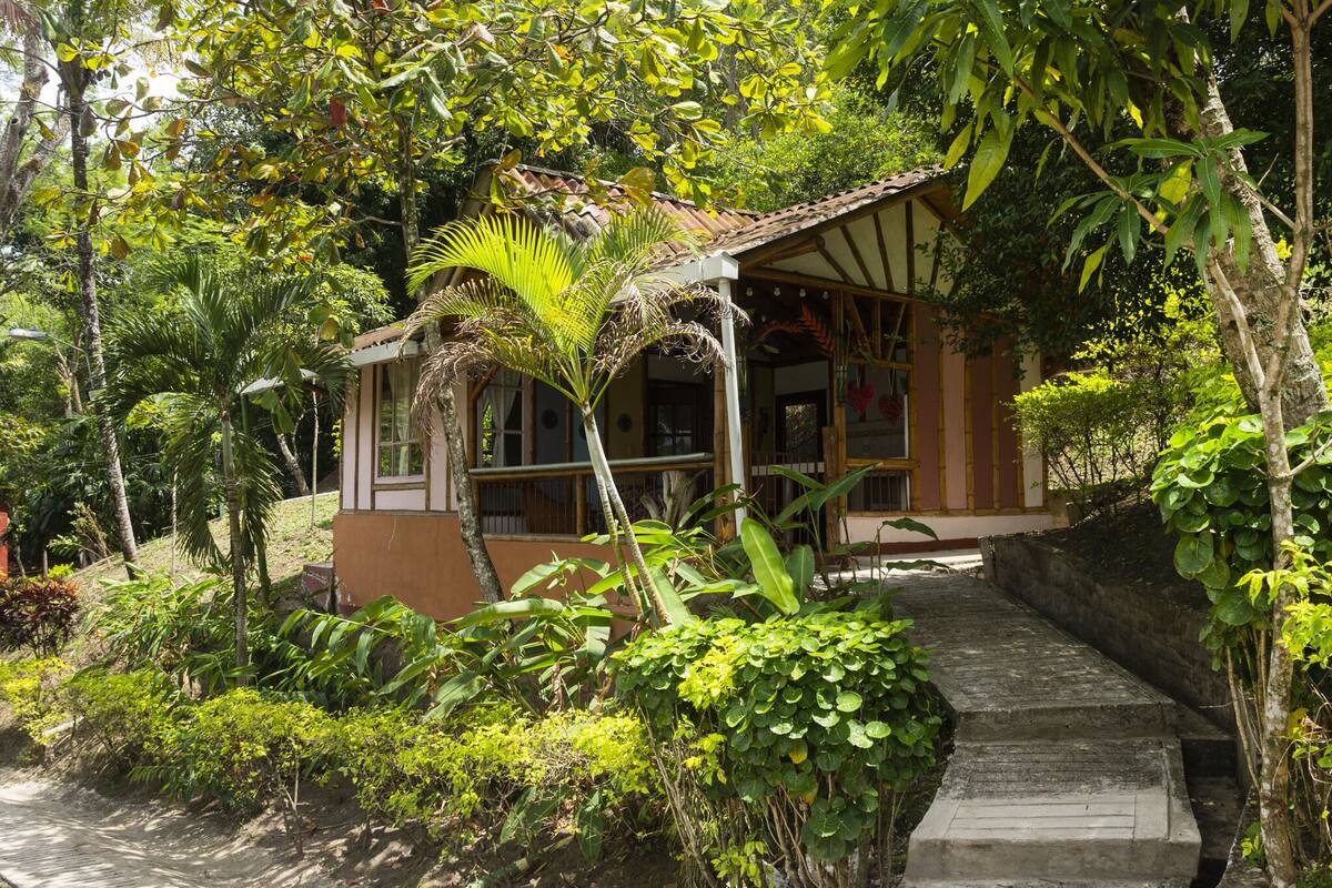 A small, rustic building with a tiled roof, surrounded by lush greenery and a stone staircase leading up to it.