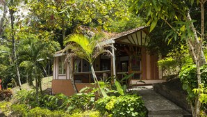 Um pequeno edifício rústico com telhado de telhas, rodeado por vegetação exuberante e uma escadaria de pedra que leva até ele.