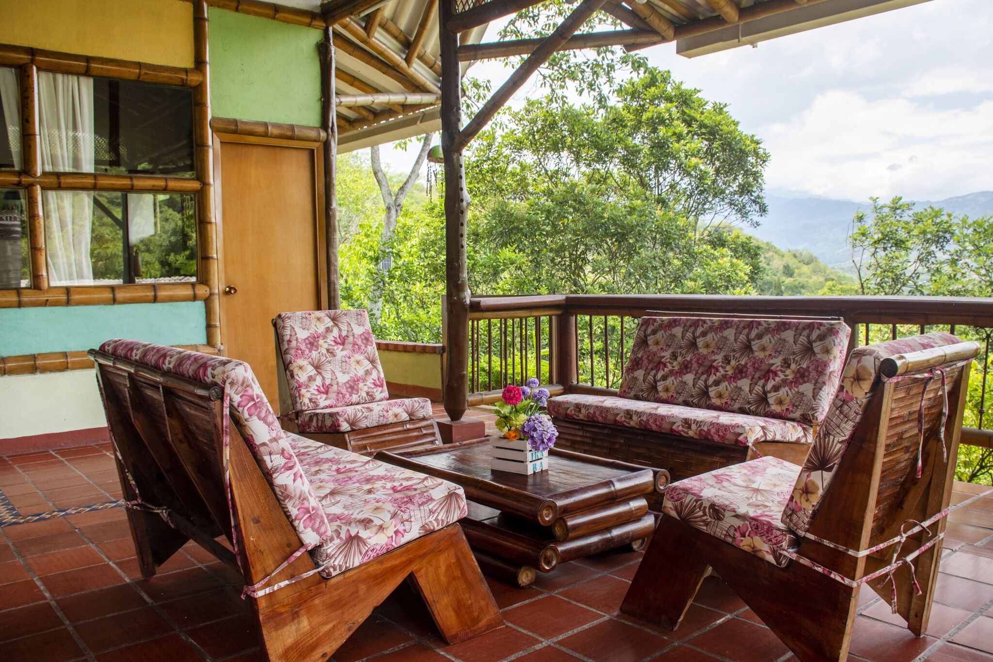Una terraza de madera con una mesa y sillas, rodeada de abundante vegetación y un telón de fondo montañoso.
