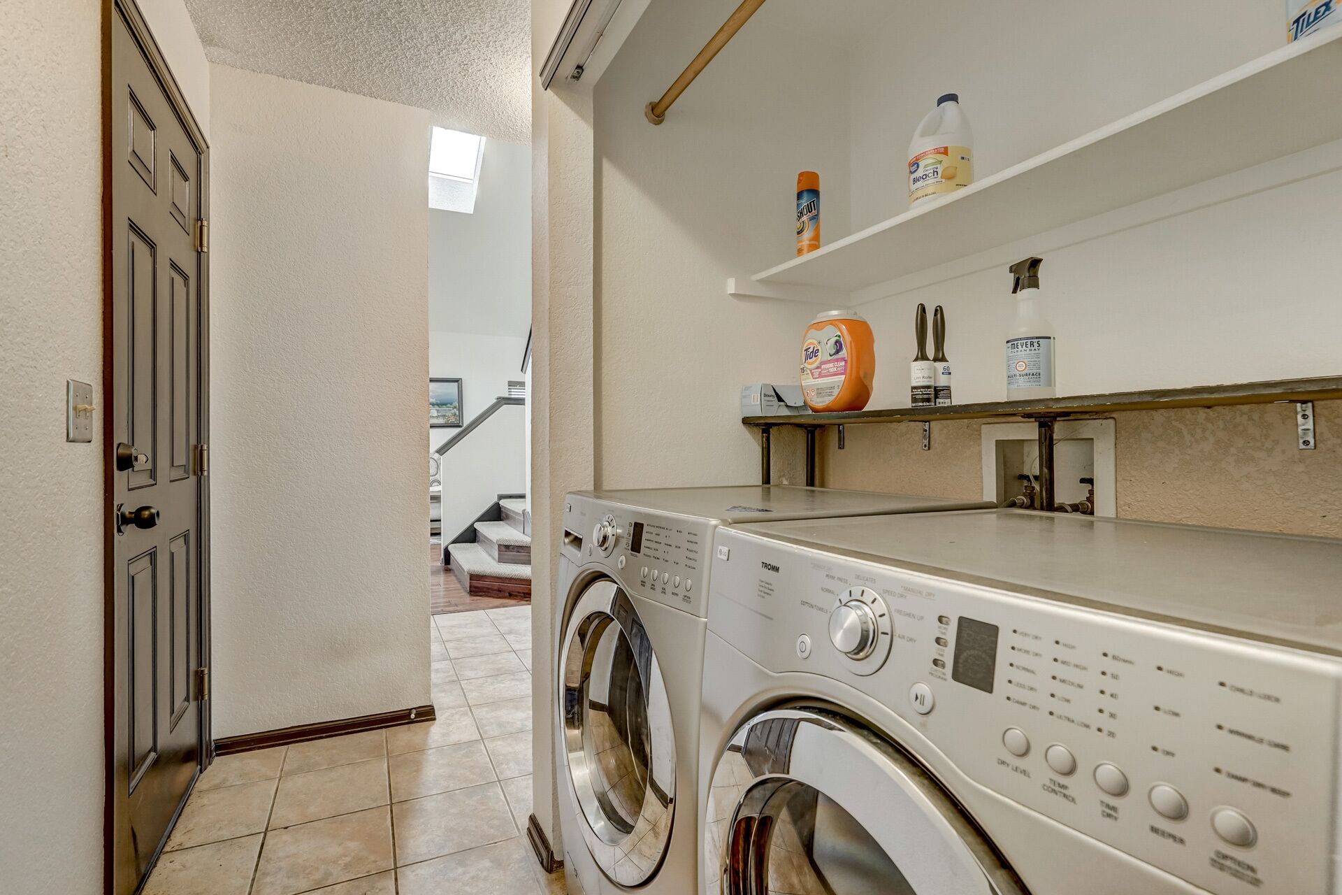 A laundry room with a front-loading washing machine, a shelf with cleaning supplies, and a door leading to another room.