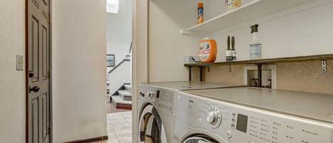 A laundry room with a front-loading washing machine, a shelf with cleaning supplies, and a door leading to another room.