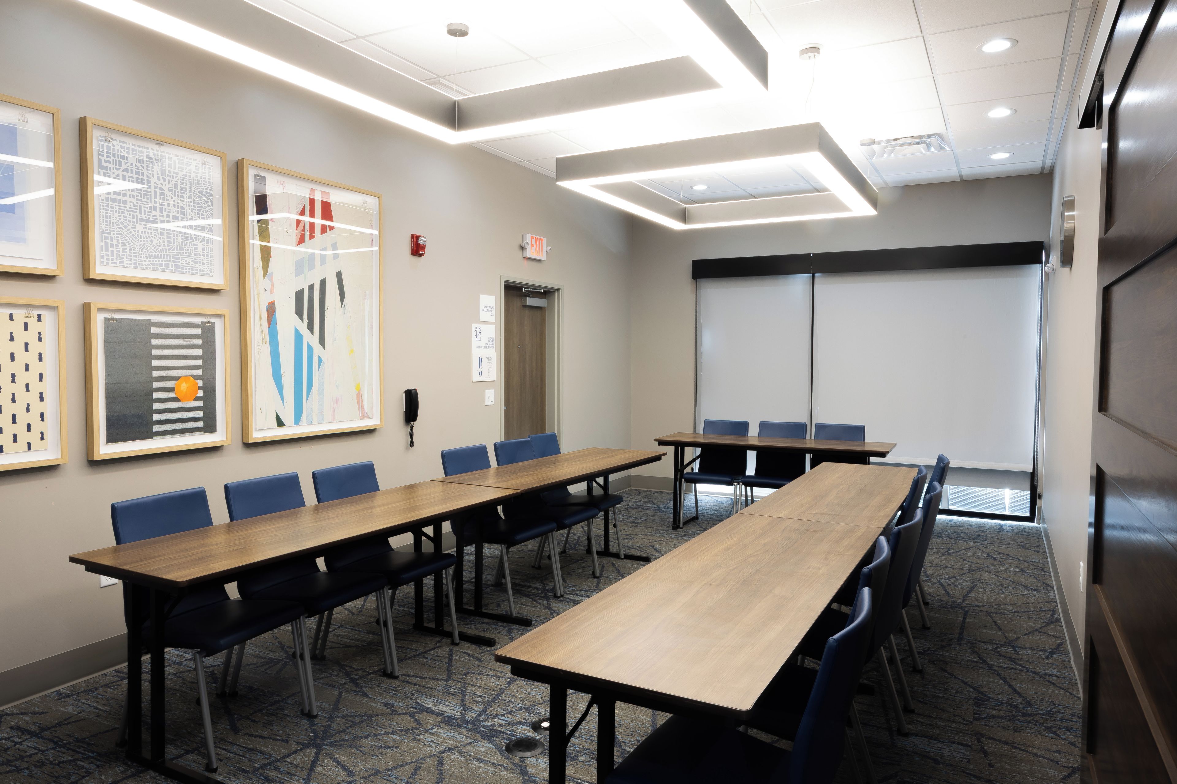 A conference room with a long table, blue chairs, framed artwork on the wall, and an exit sign.