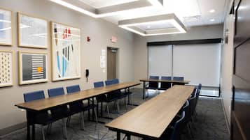 A conference room with a long table, blue chairs, framed artwork on the wall, and an exit sign.
