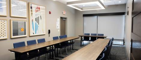 A conference room with a long table, blue chairs, framed artwork on the wall, and an exit sign.