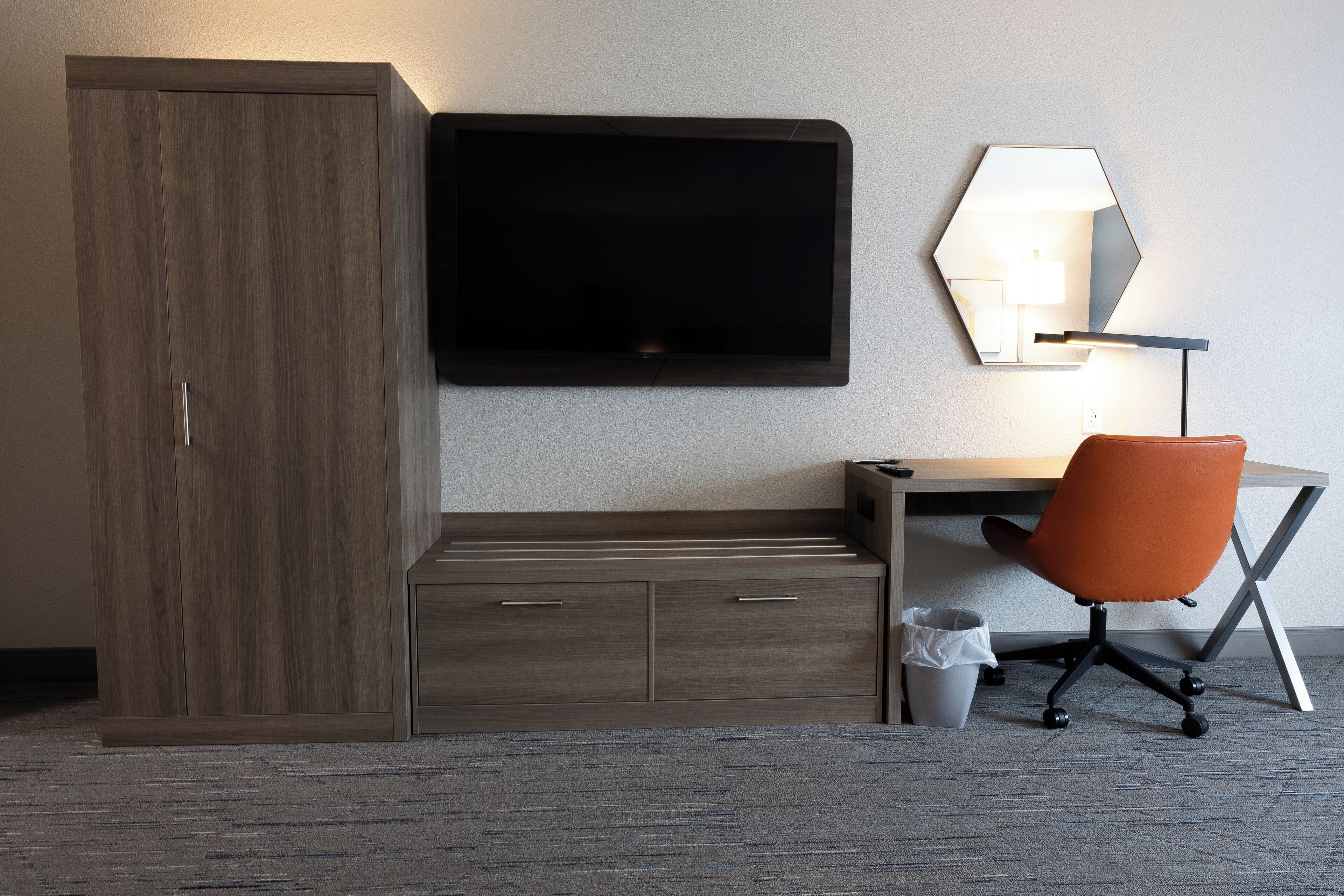 A hotel room with a wooden wardrobe, a flat-screen TV, a desk with a chair, and a wall-mounted lamp.