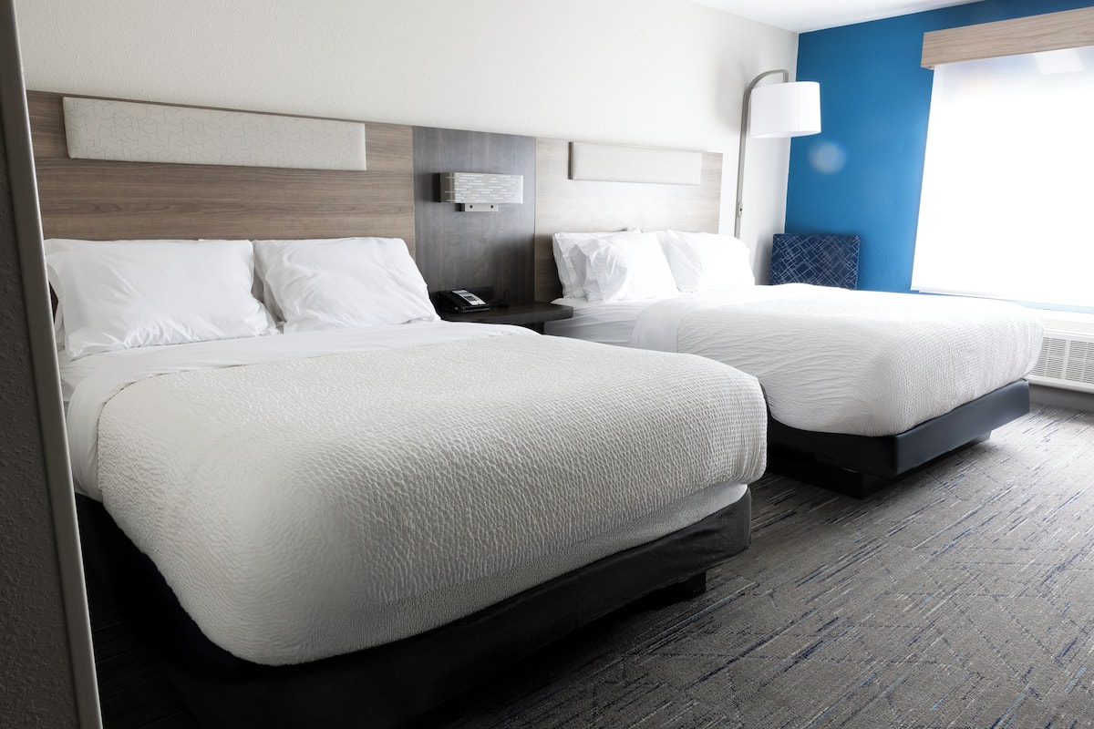 Een hotelkamer met twee bedden, een nachtkastje, een wandlamp en een raam met jaloezieën.
