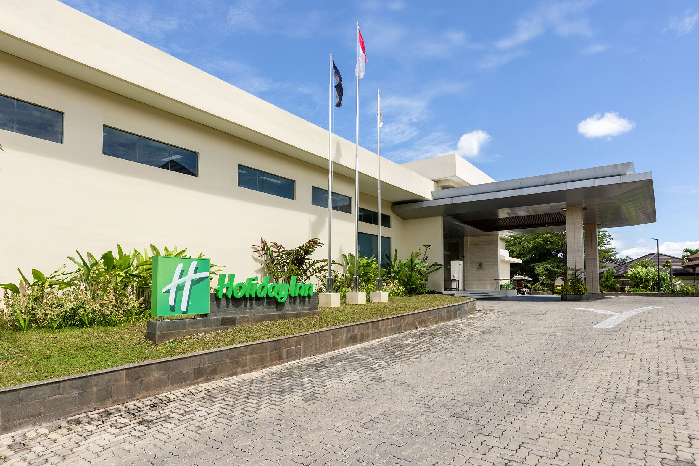 A modern building with a sign reading 'Holiday Inn,' a flagpole with flags, and a paved entrance area with greenery.