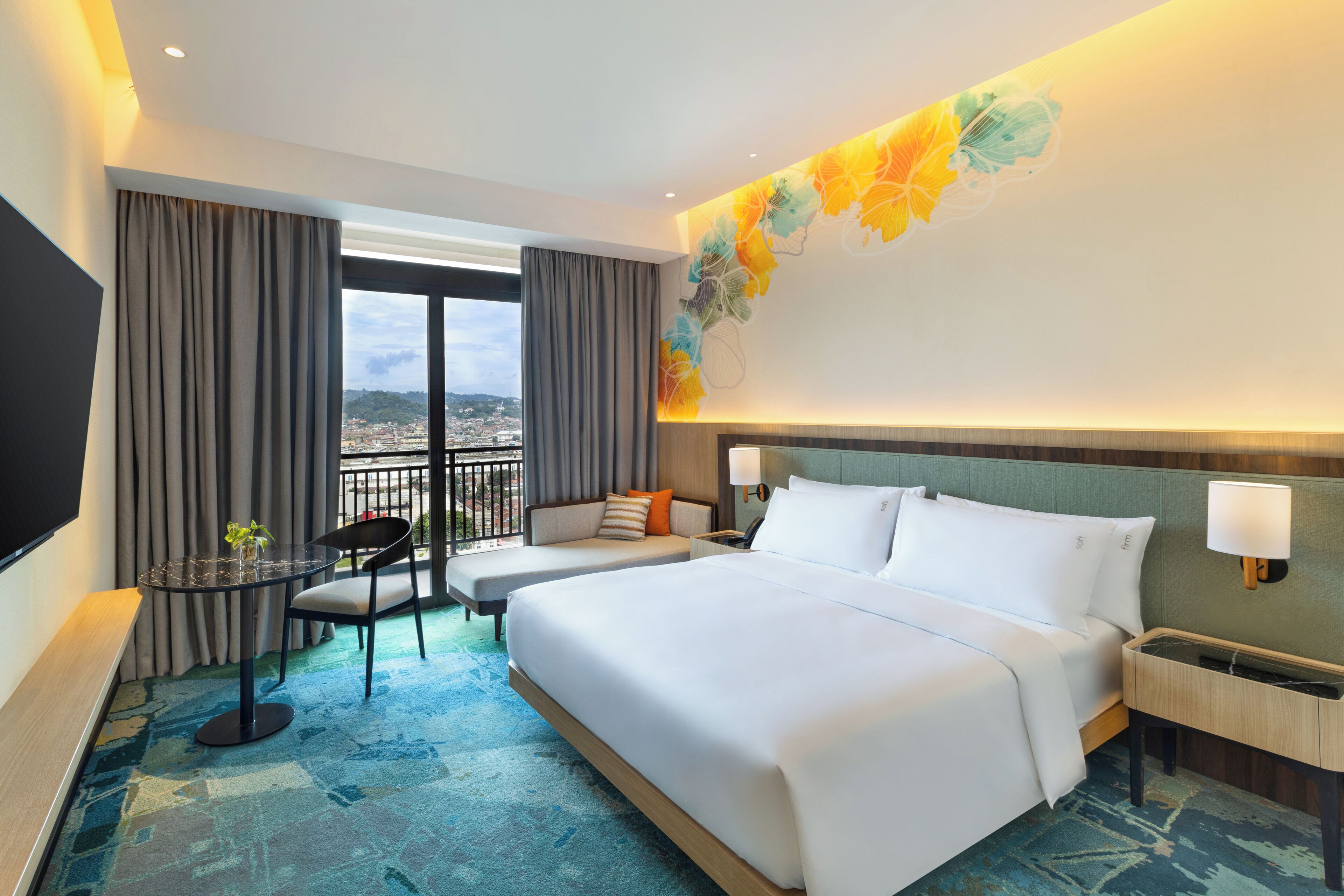 A hotel room with a large bed, a small table with a chair, a sofa, a TV, and a balcony with curtains.