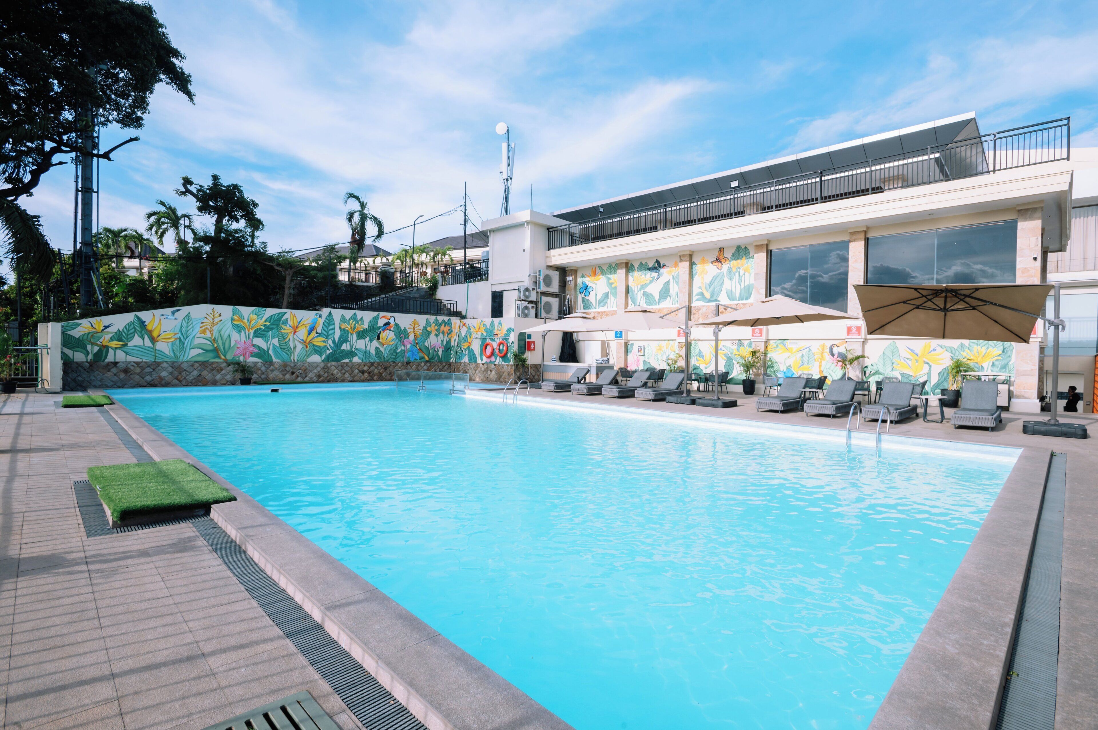 A swimming pool with lounge chairs and umbrellas, surrounded by a building with large windows and a mural.