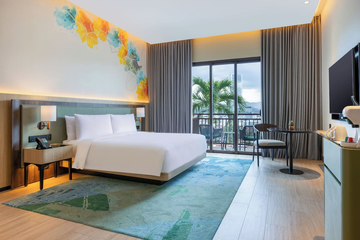 A hotel room with a large bed, a desk with a chair, a television, and a balcony with a view of palm trees and buildings.