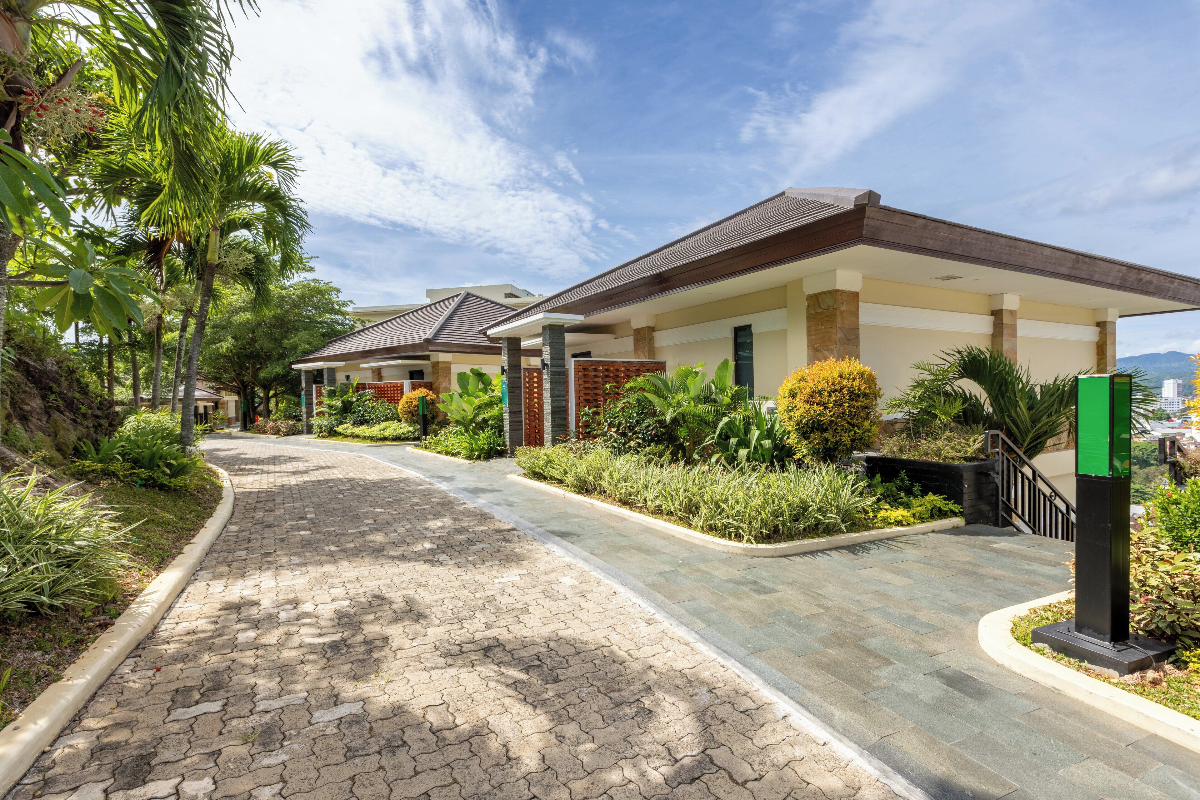A paved pathway leading to a building with a covered entrance, surrounded by lush greenery and palm trees.