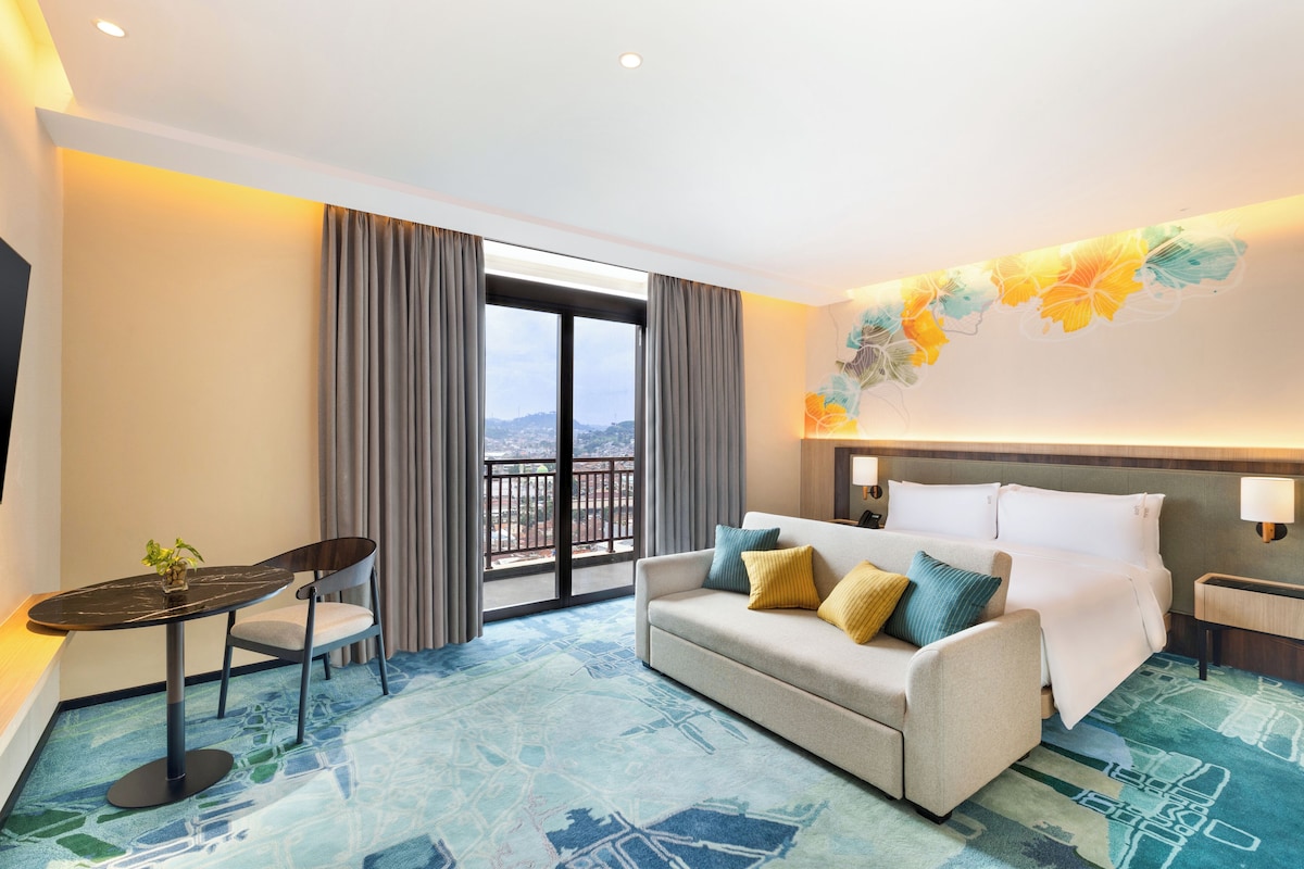 A hotel room with a sofa, a small table, a chair, a bed, a nightstand, and a large window with curtains.
