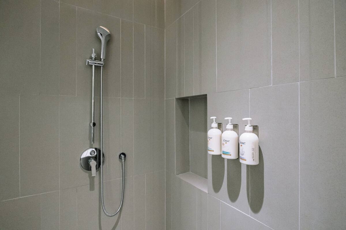A shower area with a handheld showerhead, a wall-mounted faucet, and a shelf with three bottles of Dove body wash.