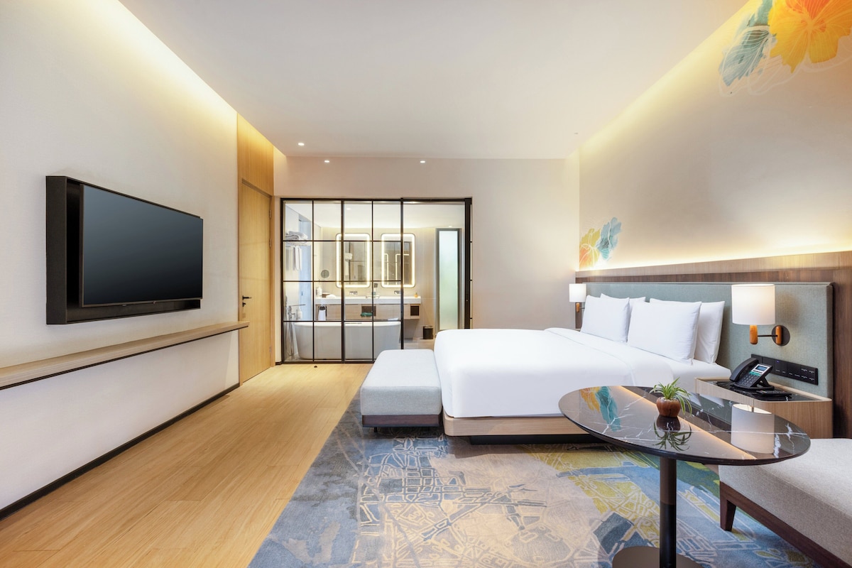 A modern hotel room with a flat-screen TV, a large bed, a small table, and a bathroom visible through a glass door.