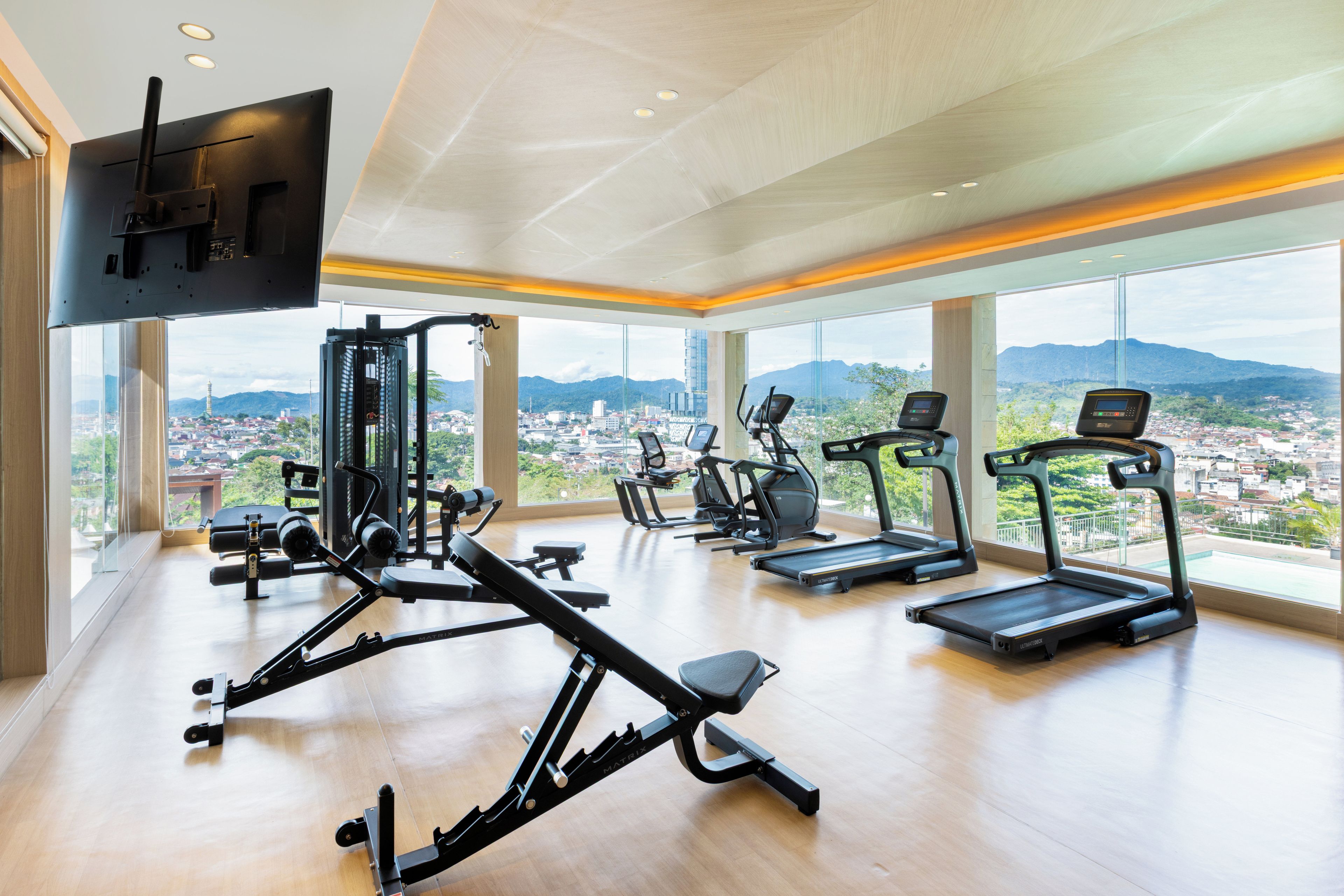 Un gimnasio con diversos equipos de ejercicio, amplias ventanas con vistas a la ciudad y un televisor de pantalla plana montado en la pared.