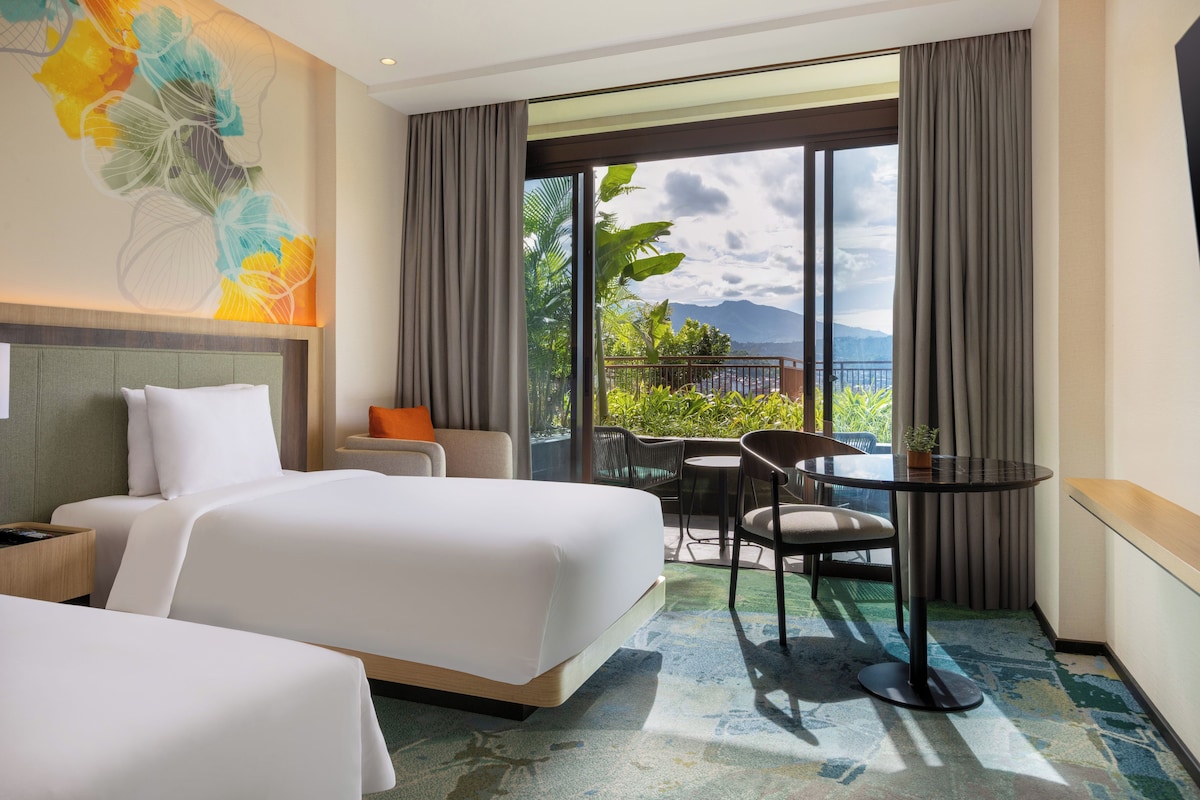 A hotel room with two beds, a seating area with a table, and a large window with a view of mountains and greenery.