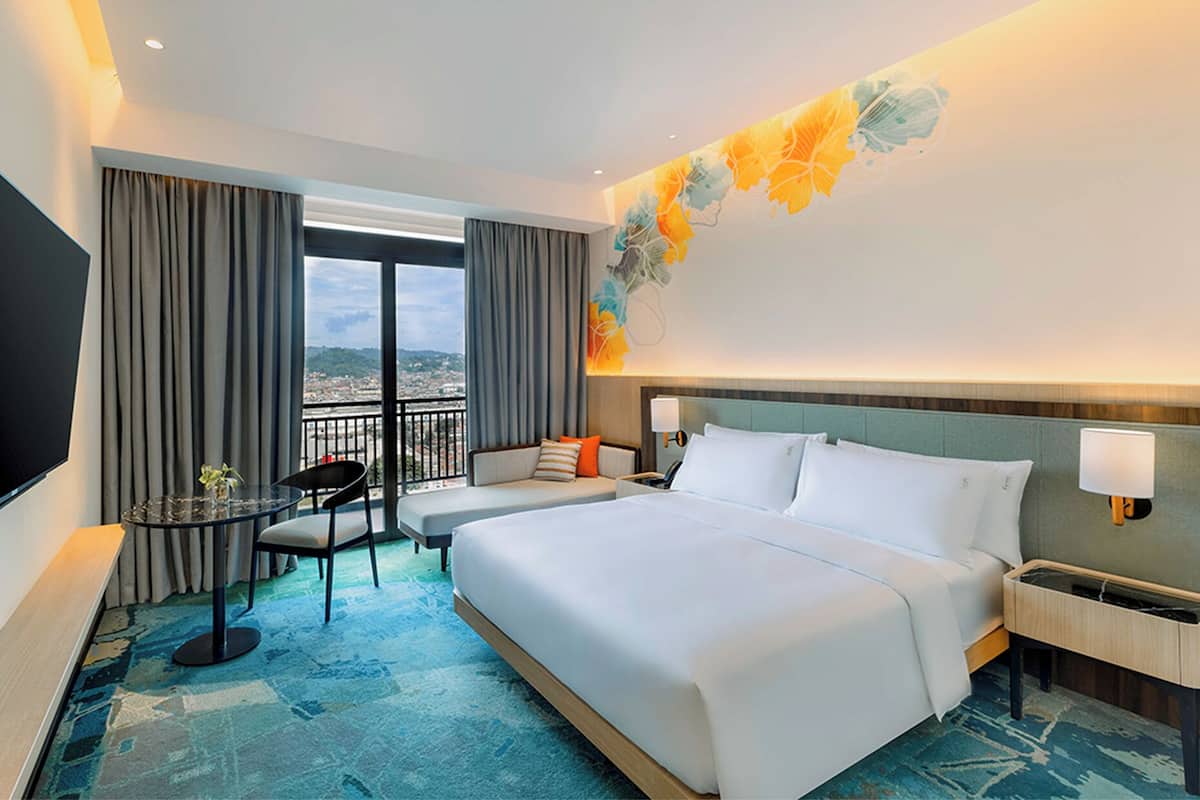 A hotel room with a large bed, a desk, a chair, and a view of the city.