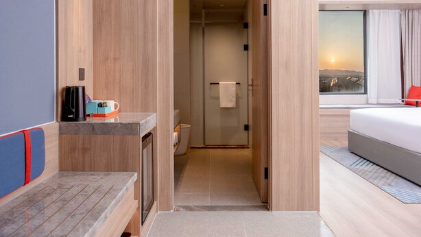 A modern hotel room with a wooden floor, a bed with a red pillow, a shower area, and a view of the city at sunset. - Holiday Inn Express Suzhou Shihu University Town by IHG (Suzhou)