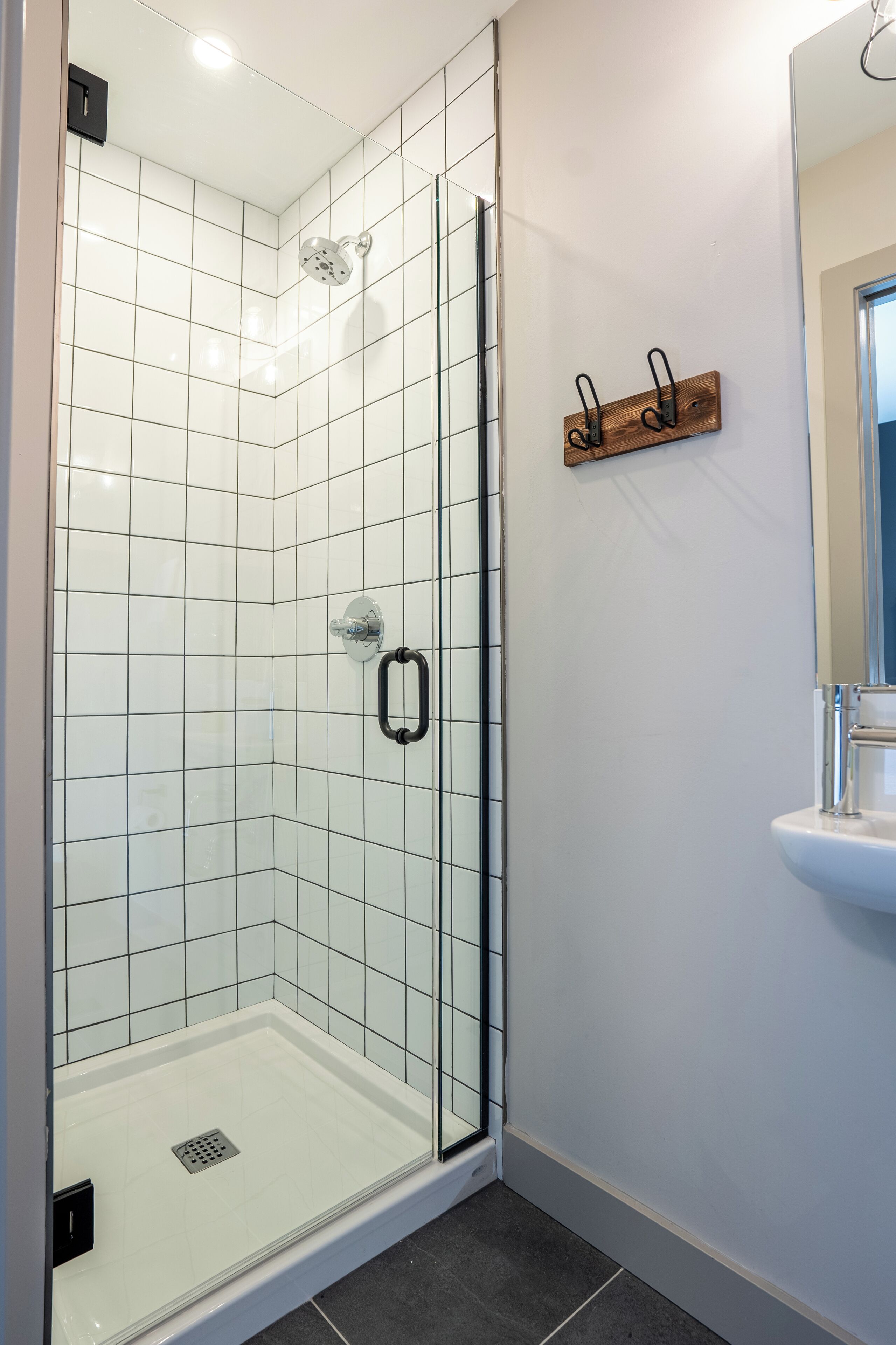 A glass-enclosed shower with white tiles, a black handle, and a wooden shelf with hooks.
