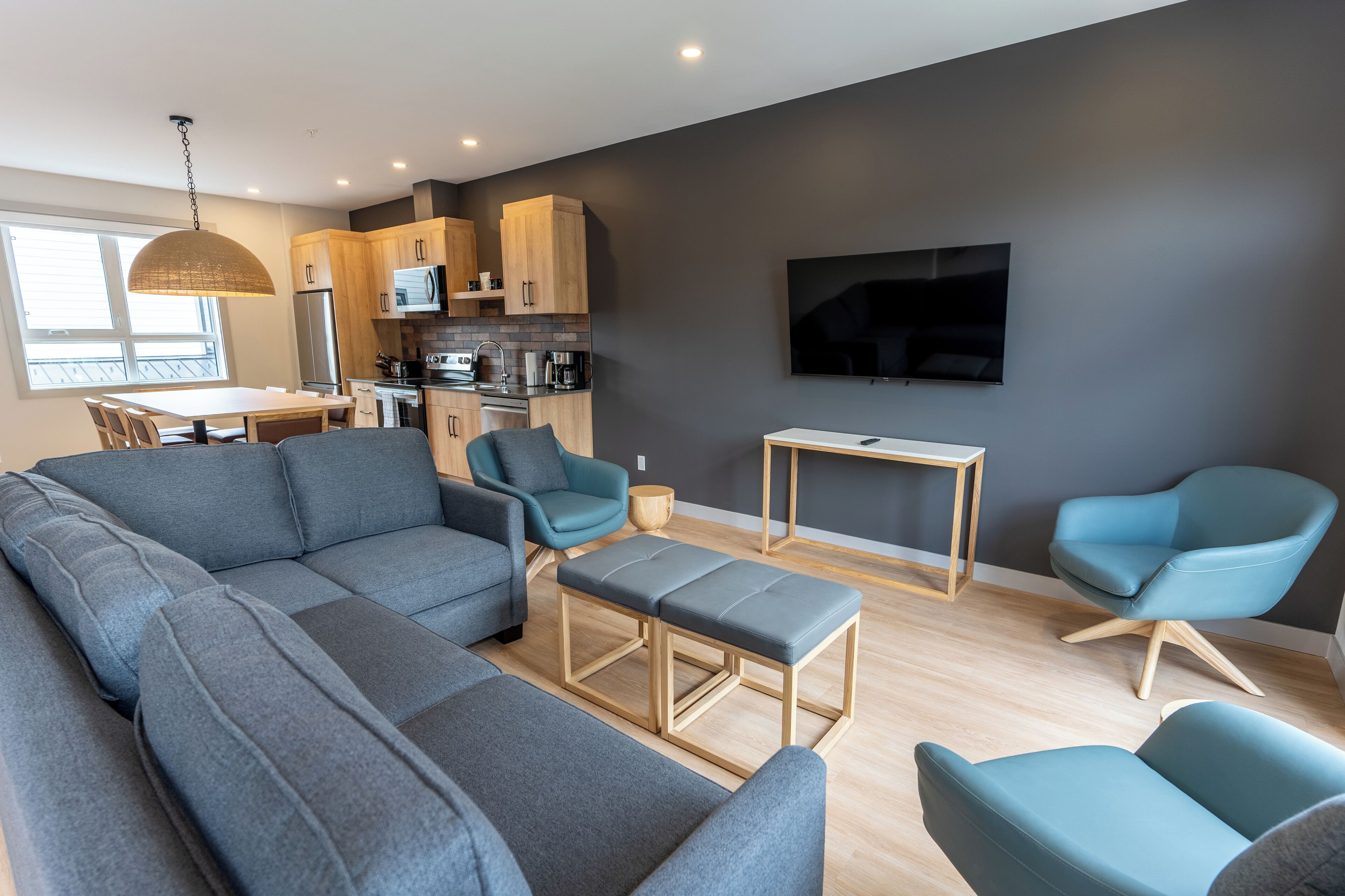 Un salon moderne avec un canapé d’angle, une table basse et deux fauteuils décoratifs. On aperçoit une cuisine avec des meubles en bois et un téléviseur à écran plat à l’arrière-plan.