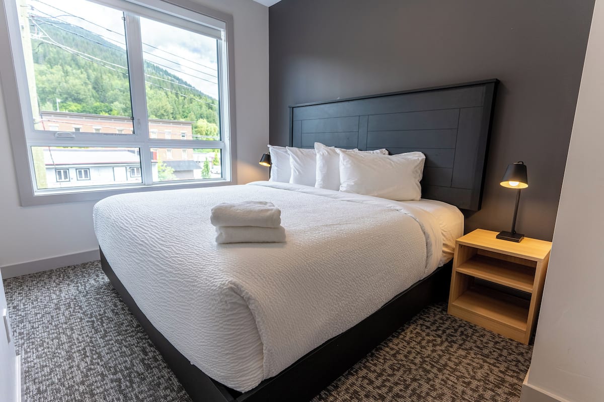 A neatly made bed with white linens and a folded towel, a dark headboard, a bedside table with a lamp, and a window with a view of buildings and greenery.