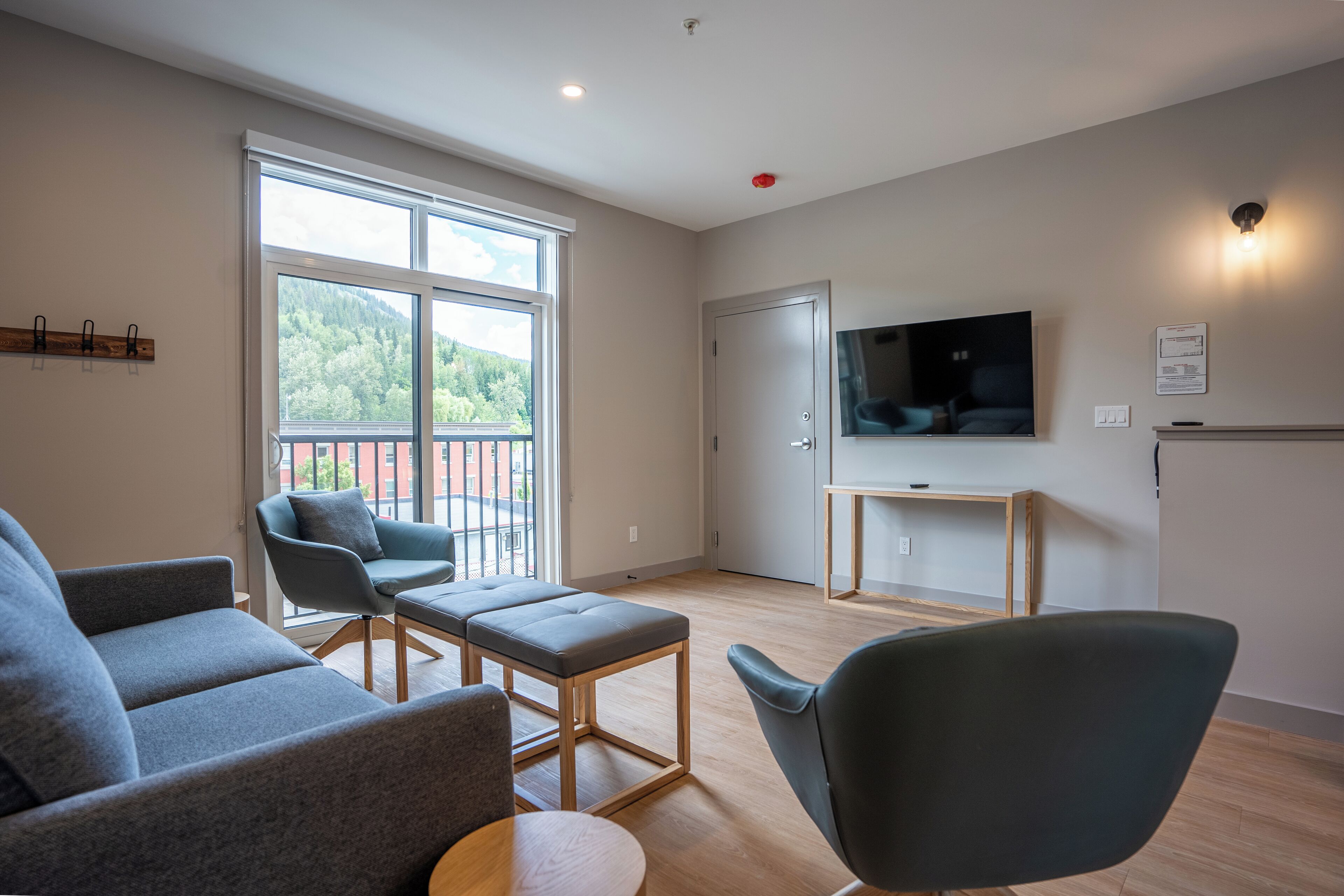 Een moderne woonkamer met een bank, fauteuil, poef, een televisie en een balkon met uitzicht op groen.