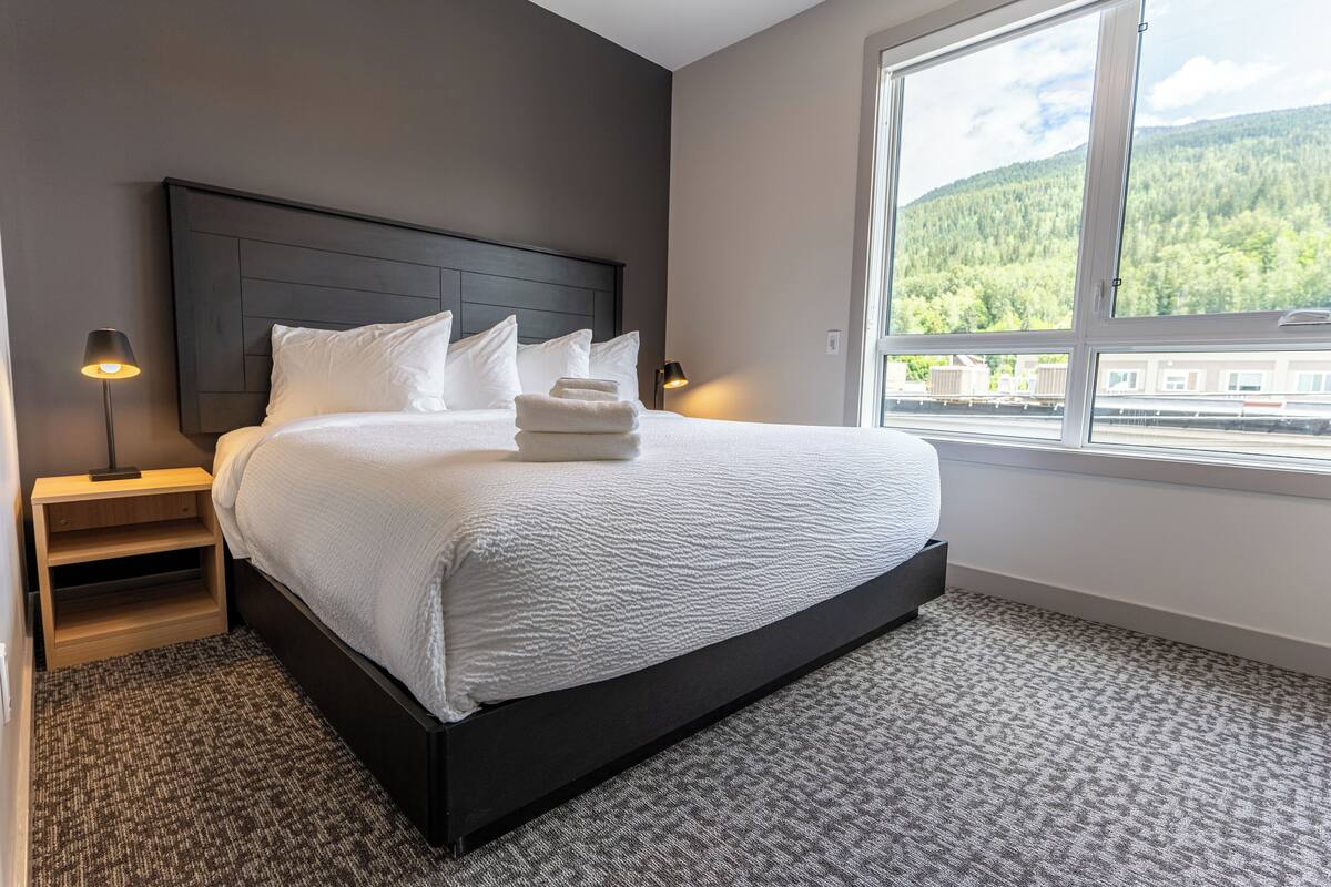 A modern hotel room with a large bed, a nightstand, a lamp, and a window with a mountain view.
