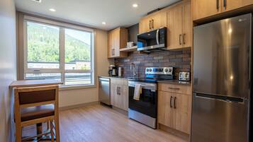 A modern kitchen with wooden cabinets, stainless steel appliances, and a large window offering a view of a forested area.