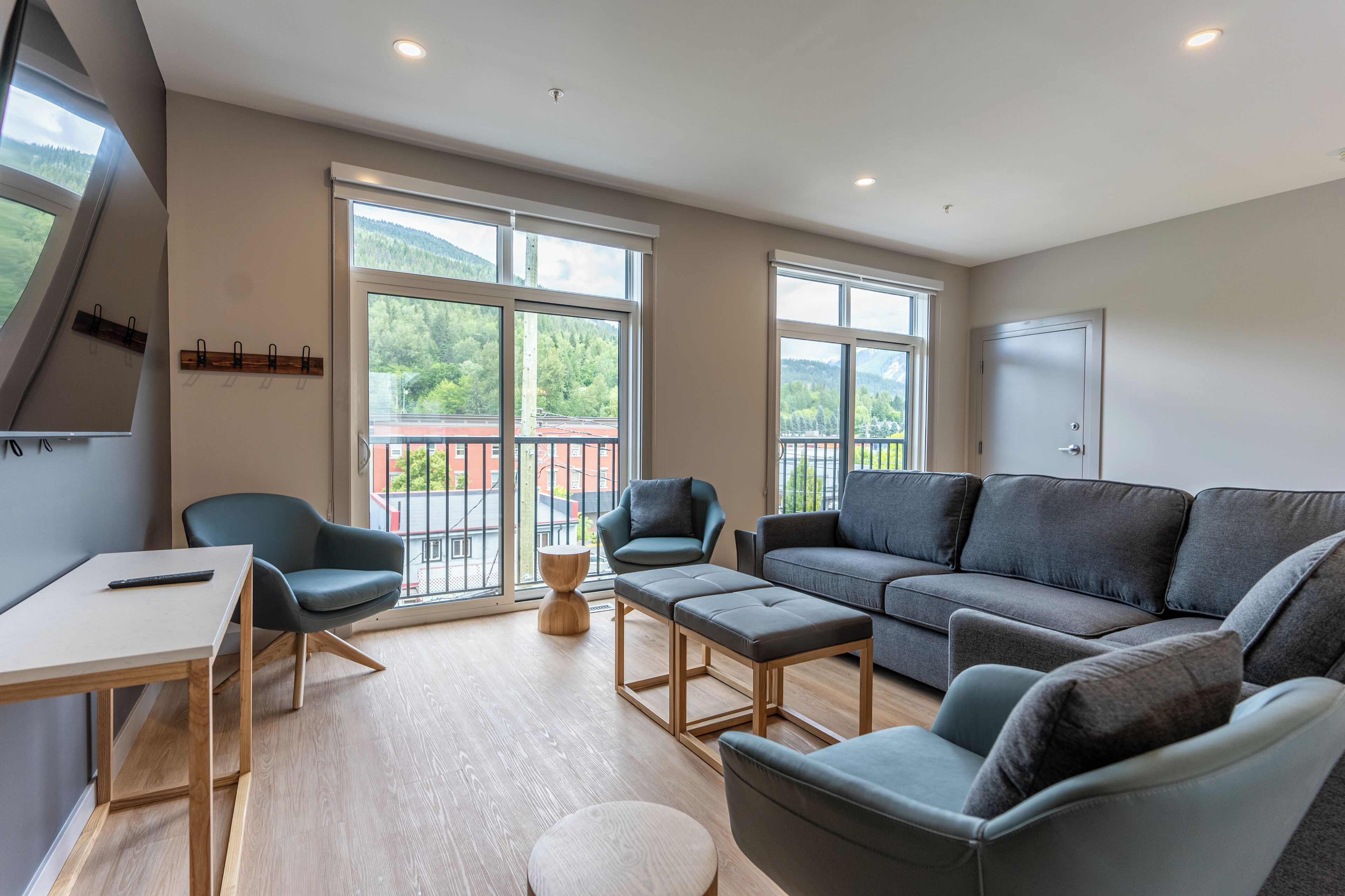 Un salon moderne comprenant un canapé, des fauteuils, une table basse et un téléviseur. De grandes fenêtres offrent une vue sur la verdure et donnent accès à un balcon.