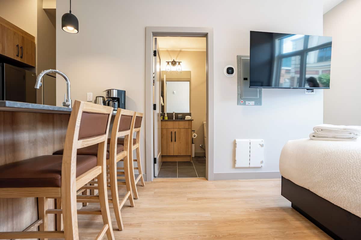 A modern hotel room with a wooden dining table, chairs, a bed, a TV, and a bathroom visible through an open door.