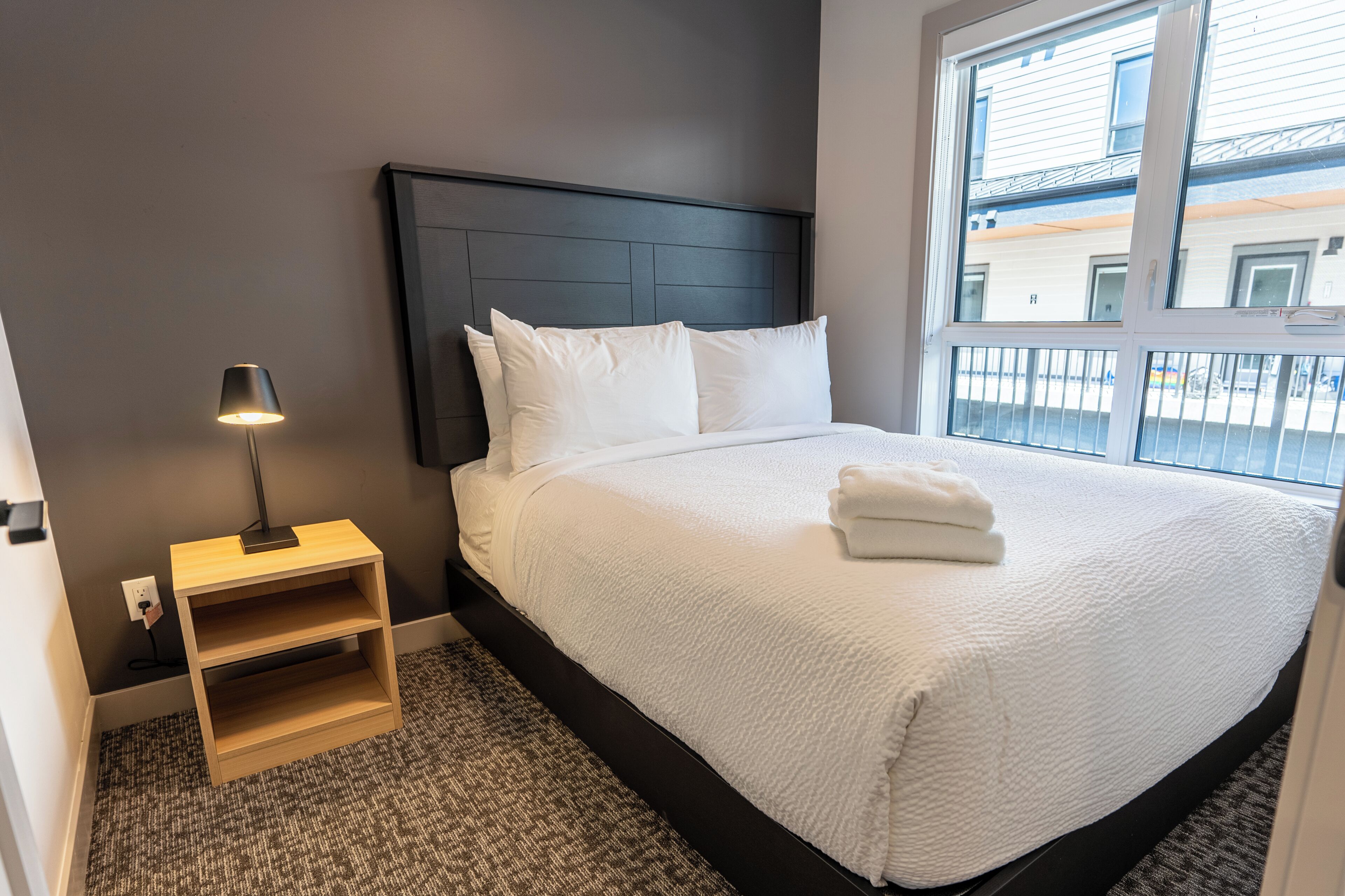 A neatly made bed with white linens and pillows, a bedside table with a lamp, and a view of buildings through a window.
