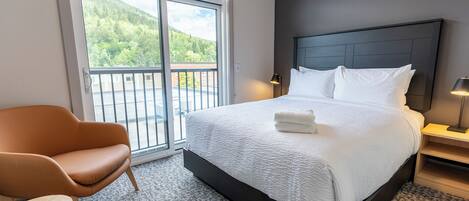A hotel room with a large bed, a chair, a small table, and a view of a balcony with a mountain backdrop.