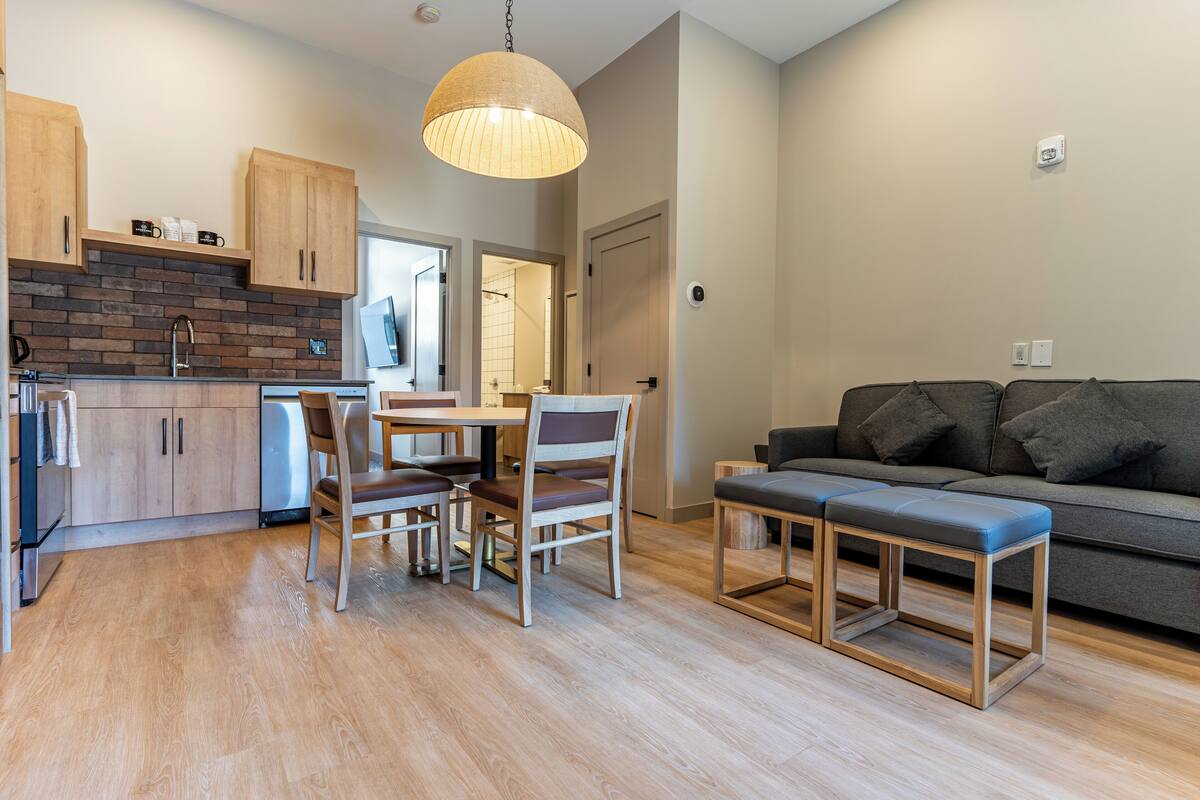 A modern kitchen with wooden cabinets, a dining area with chairs, a bench, and a grey sofa in a living area.