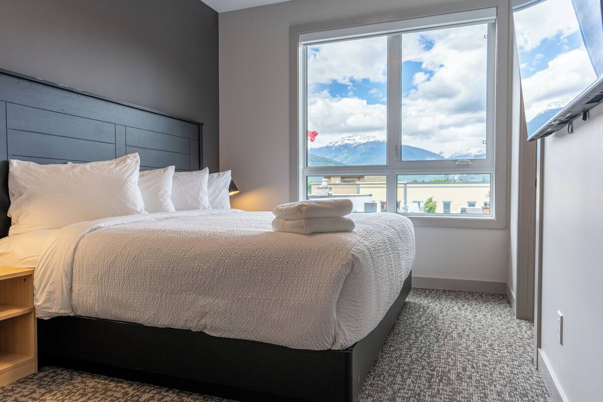 A neatly made bed with white linens and a folded towel, a window with a view of mountains, and a dark headboard.