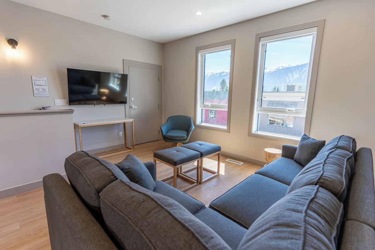 A modern living room with a large grey sofa, a small blue chair, a TV on a stand, and large windows with a mountain view.