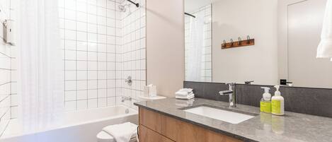 Une salle de bain moderne avec une baignoire, des toilettes et un lavabo. On peut voir deux bouteilles de shampoing et d’après-shampoing sur le lavabo.