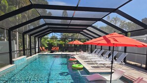 Piscina techada con sillas de descanso y sombrillas, rodeada por una valla y árboles.