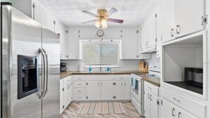 Una cocina moderna con armarios blancos, electrodomésticos de acero inoxidable, una ventana con persianas y un ventilador de techo.