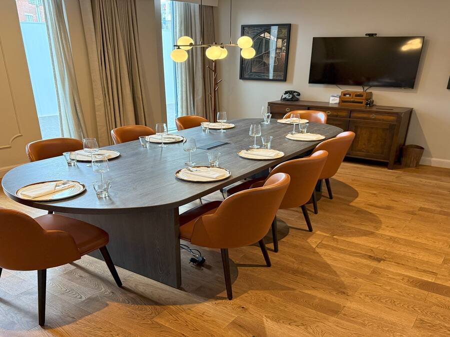 Un comedor con una mesa larga dispuesta para seis personas, rodeada por seis sillas naranjas. Hay un televisor montado en la pared y un mueble a la derecha.