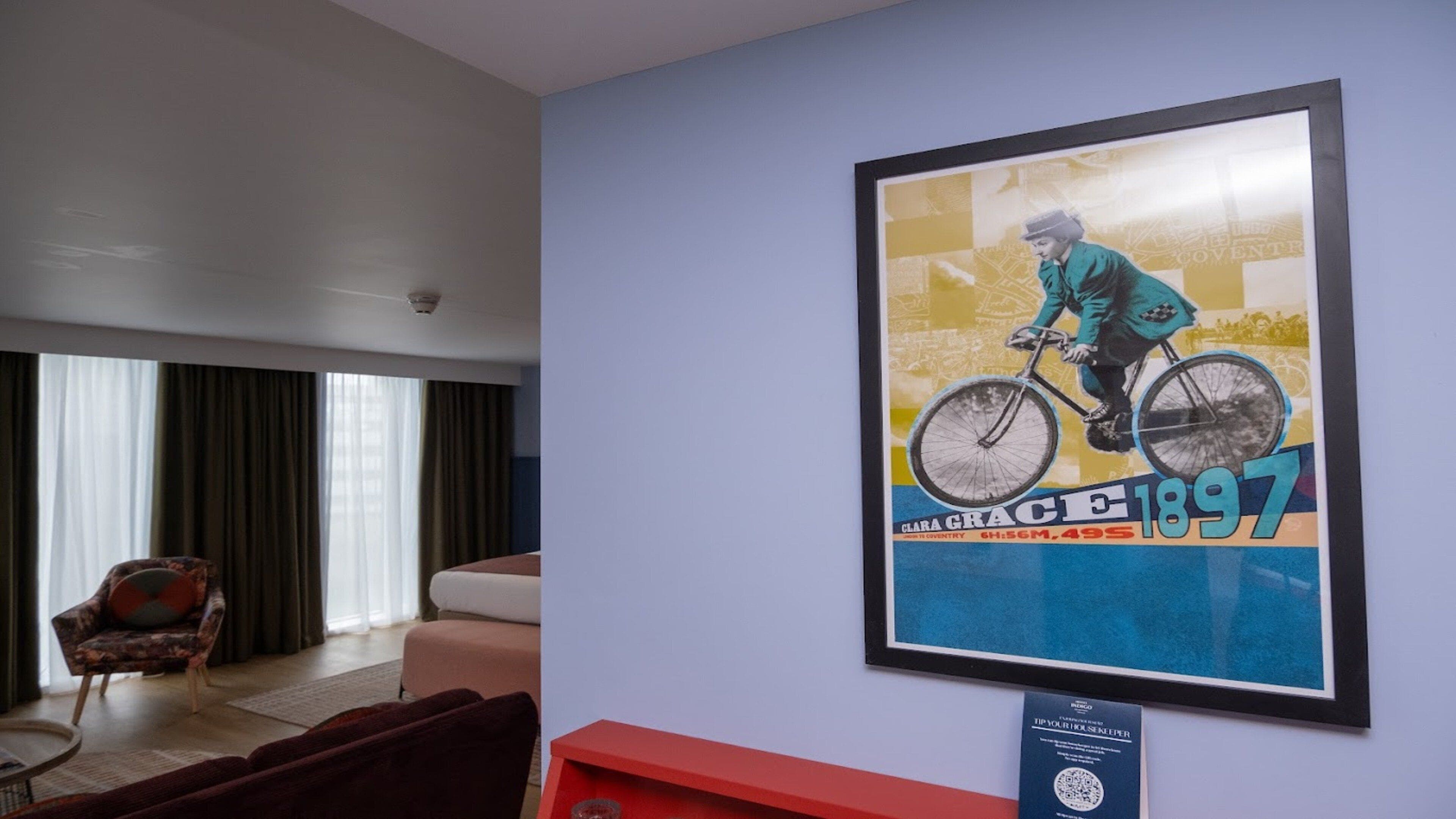 A hotel lobby with a framed poster of a cyclist, a red side table, and a patterned armchair.