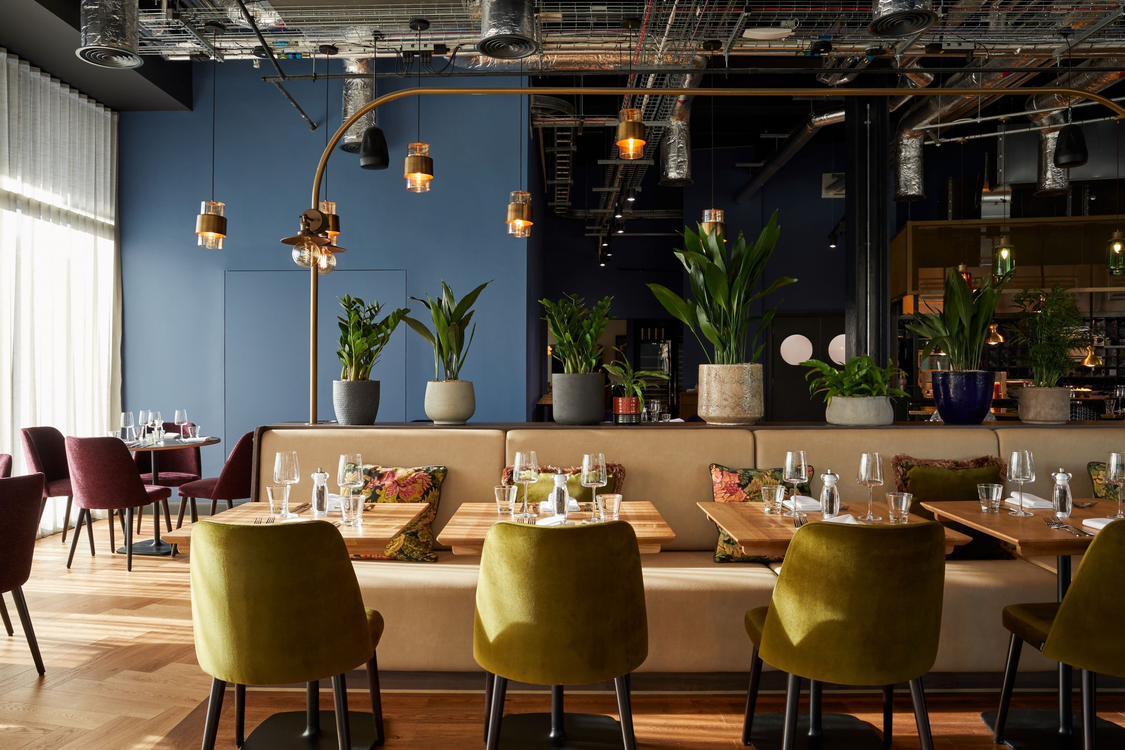 A modern restaurant with wooden tables and green chairs, potted plants, and hanging pendant lights.
