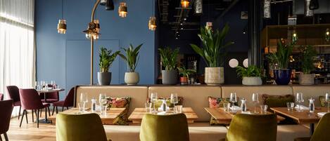 A modern restaurant with wooden tables and green chairs, potted plants, and hanging pendant lights.