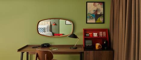Habitación de hotel con un escritorio de madera, una silla, teléfono, lámpara, estantería con aperitivos y vasos, y un cuadro en la pared.