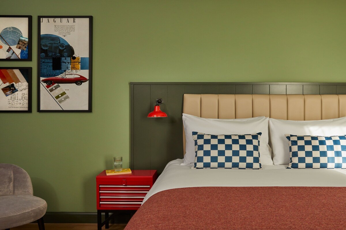A bedroom with a bed, two checkered pillows, a red bedside table with a glass, and a red lamp.