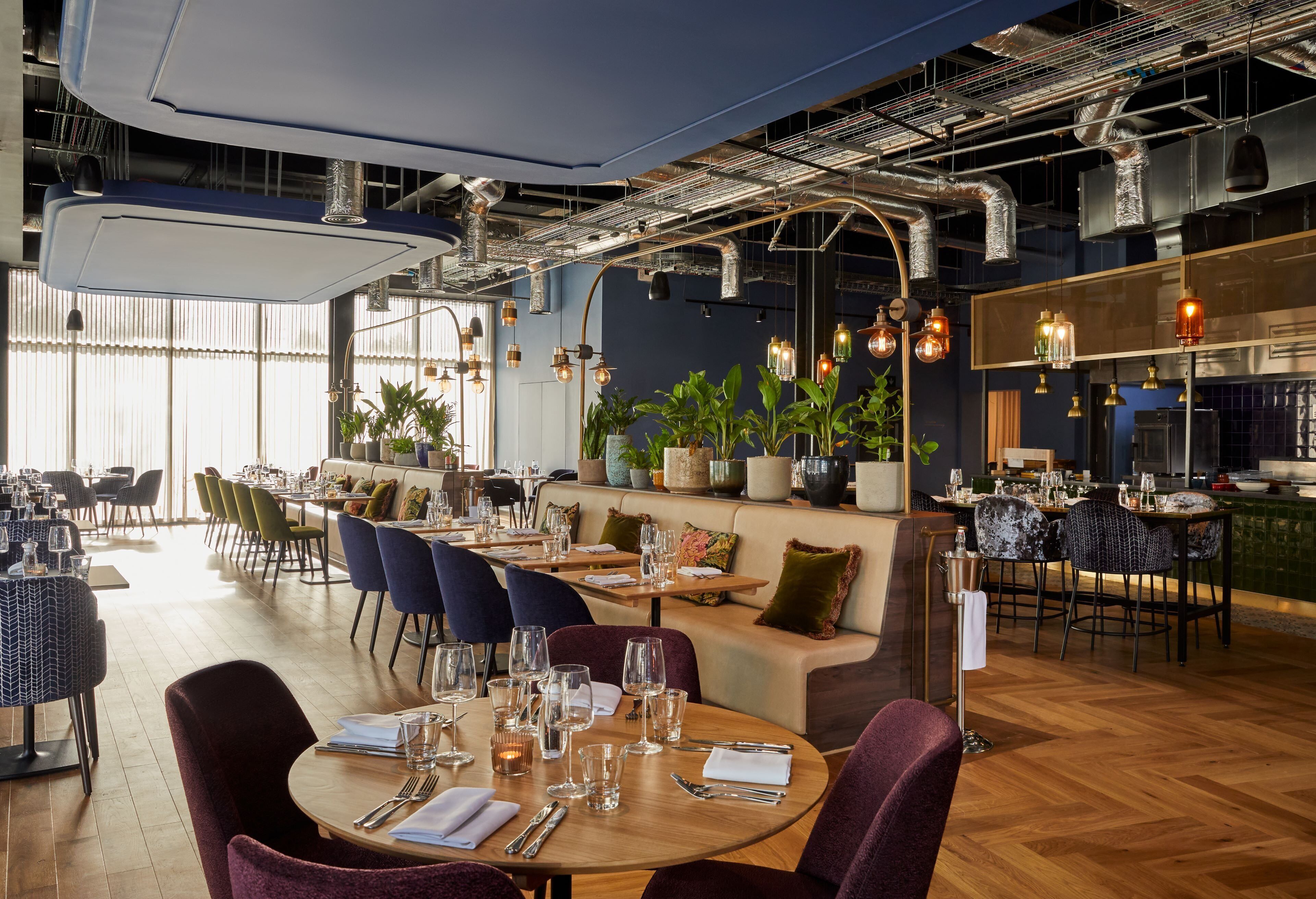 A modern restaurant with wooden tables, cushioned seating, and hanging pendant lights.