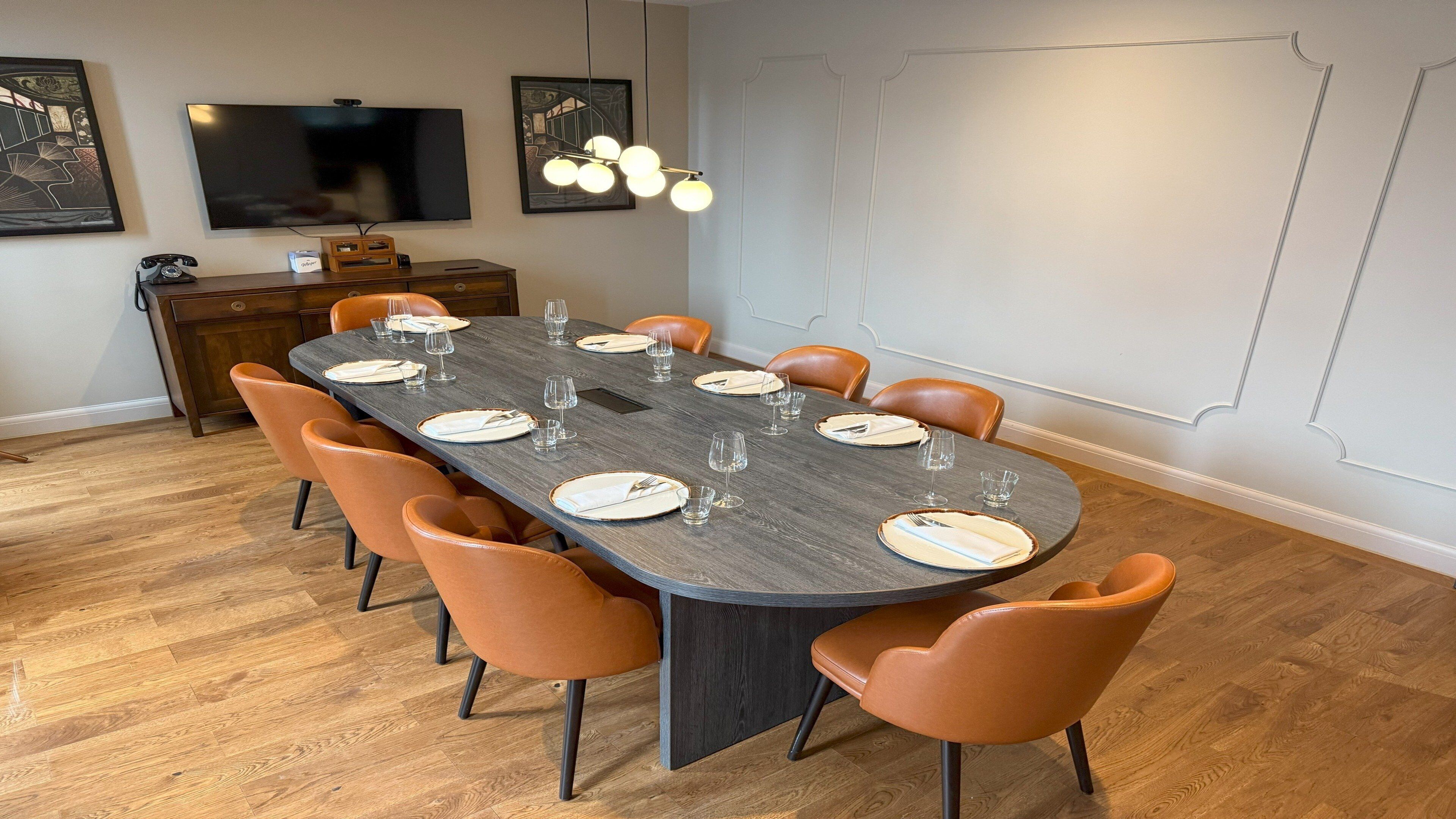 A dining area with a long table set for six, surrounded by orange chairs, a wooden cabinet, and a flat-screen TV on the wall.