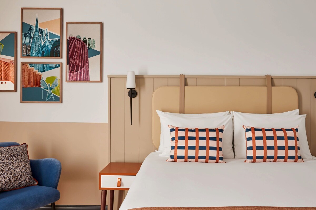 A hotel room with a bed, two pillows, a nightstand, a lamp, a chair, and four framed artworks on the wall.