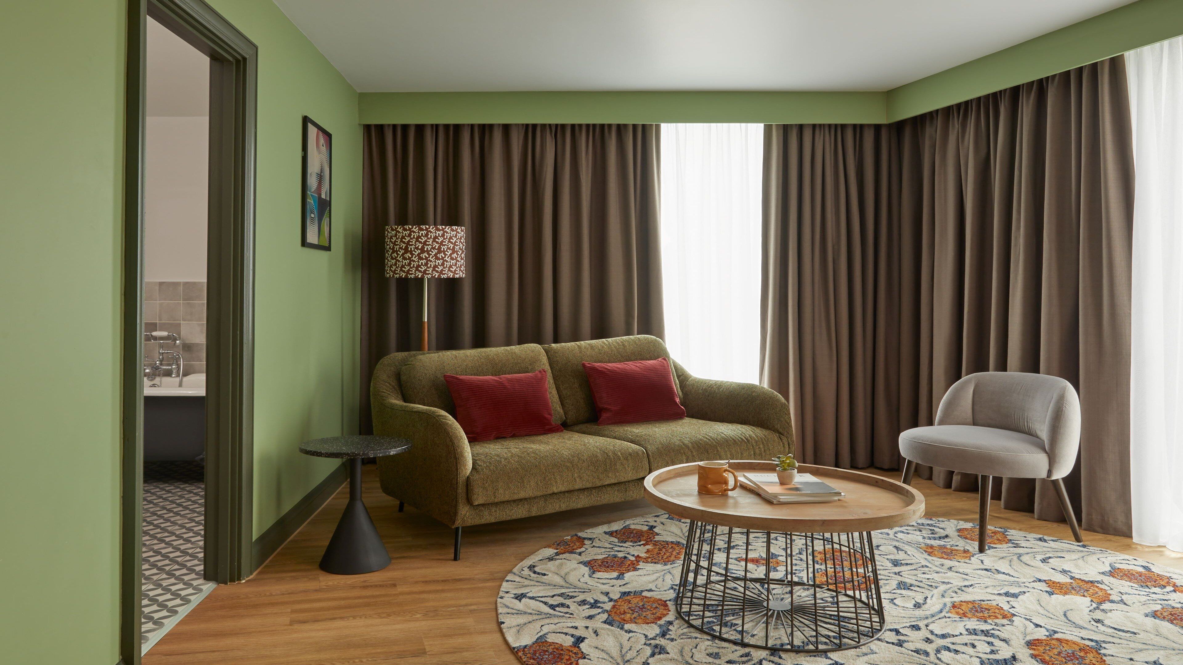 A living room with a green sofa, red cushions, a round coffee table, a grey armchair, a patterned rug, and a floor lamp.